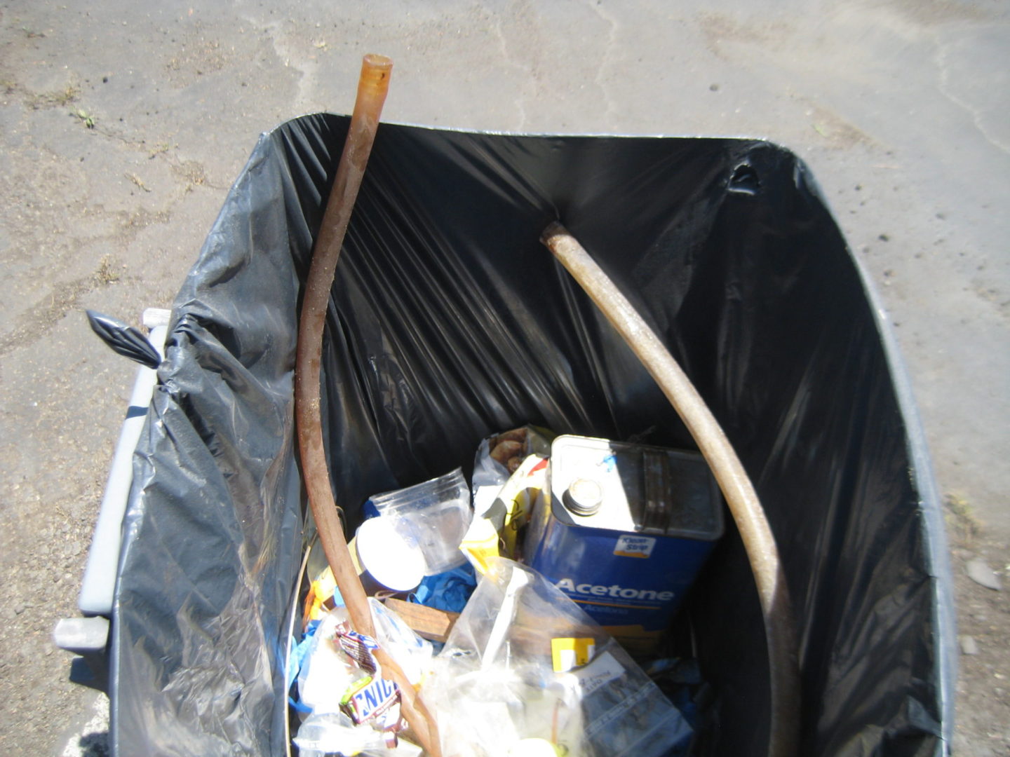 A trash can filled with garbage next to a wooden stick.